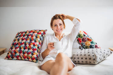 Junge Frau mit Nachthemd sitzt morgens im Haus auf dem Bett und hält eine Tasse Kaffee in der Hand. - HPIF22853