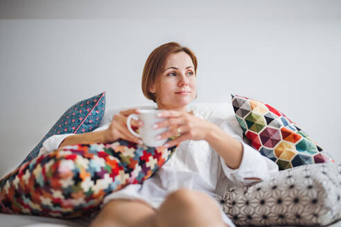 Junge Frau mit Nachthemd sitzt morgens im Haus auf dem Bett und hält eine Tasse Kaffee in der Hand. - HPIF22855