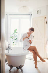 A young woman in the morning in a bathroom sitting on a bath tub, applying nail polish. - HPIF22880