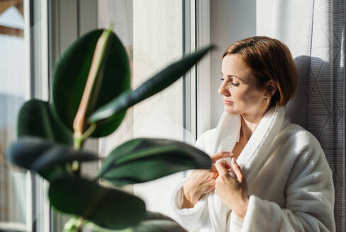 A young woman with bath robe and closed eyes standing indoors by a window in the morning, meditating. Copy space. - HPIF22884
