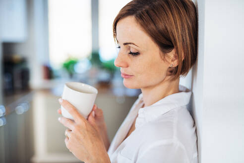 Eine junge, zufriedene Frau mit einer Tasse Kaffee, die in einem Haus steht und sich an eine Wand lehnt; Raum kopieren. - HPIF22889