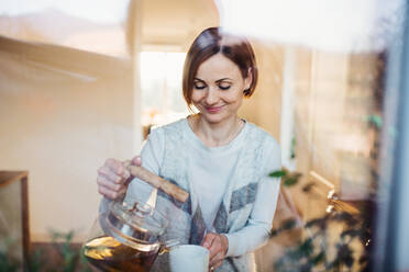 A happy young woman standing indoors in kitchen, pouring tea. Shot through glass. - HPIF22906