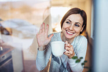 A young woman with cup of coffee looking out of a window, waving goodbye to her husband in the morning. Shot through glass. - HPIF22907