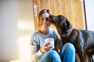 Eine junge Frau mit einer Tasse Kaffee sitzt zu Hause auf dem Boden und spielt mit einem Hund. - HPIF22910