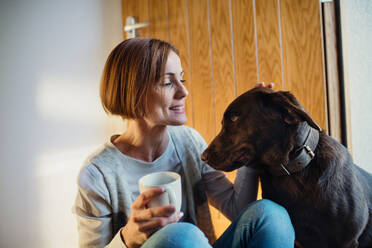 Eine junge Frau mit einer Tasse Kaffee sitzt zu Hause auf dem Boden und spielt mit einem Hund. - HPIF22911