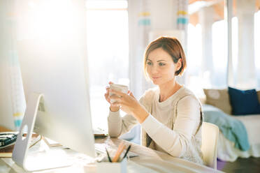 Eine glückliche junge Frau mit Smartphone, Computer und Kaffee sitzt am Schreibtisch in einem Haus und arbeitet im Home Office. - HPIF22917