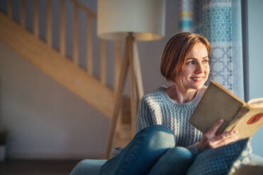 A cheerful young woman sitting indoors on a sofa at home, reading a book. Copy space. - HPIF22930