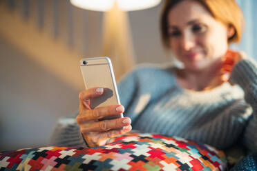 A happy young woman sitting indoors on a sofa in the evening at home, using smartphone. - HPIF22932