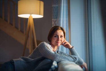 Eine glückliche junge Frau, die abends zu Hause auf einem Sofa sitzt und in die Kamera schaut. - HPIF22934