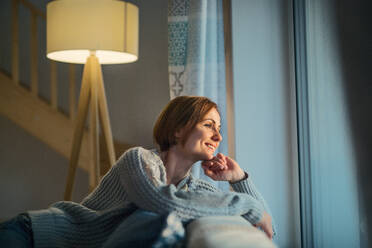 A happy young woman sitting indoors on a sofa in the evening at home, looking out of a window. - HPIF22935