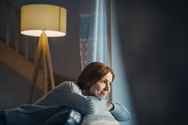 A happy young woman sitting indoors on a sofa in the evening at home, looking out of a window. - HPIF22936