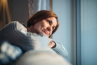 A happy young woman sitting indoors on a sofa in the evening at home, looking out of a window. - HPIF22937