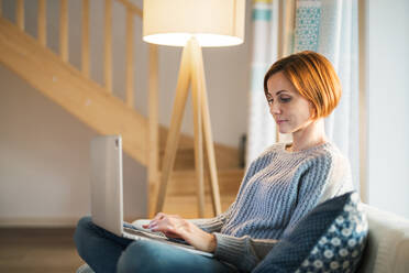 A happy young woman sitting indoors on a sofa in the evening at home, using laptop. - HPIF22938