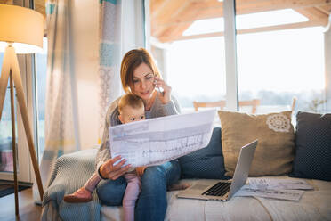 A young woman on the phone with a laptop, blueprints and toddler daughter sitting indoors, working in home office. - HPIF22941