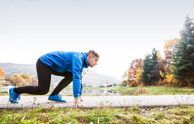 Junge Läuferin in blauer Jacke am See auf einem asphaltierten Weg, der durch grünes Gras führt, in stabiler Position. Trailrunnerin trainiert für Crosslauf draußen in bunter sonniger Herbstnatur. - HPIF23143