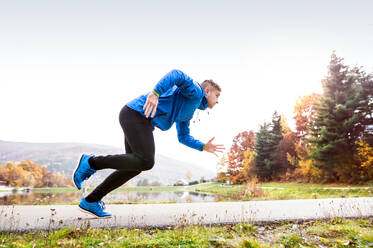 Junge Läuferin in blauer Jacke am See auf einem asphaltierten Weg. Trailrunnerin trainiert für Crosslauf draußen in bunter sonniger Herbstnatur. - HPIF23144