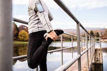 Unbekannter junger Sportler mit Smartphone in grauer Jacke, der sich im Freien streckt. Trailrunner trainiert für Crosslauf in bunter sonniger Herbstnatur. - HPIF23158