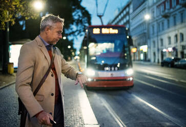 Reifer Geschäftsmann mit Koffer, der abends in Prag auf eine Straßenbahn oder einen Oberleitungswagen wartet und auf die Uhrzeit achtet. - HPIF23162