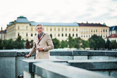 Reifer, gutaussehender Geschäftsmann mit Koffer und Smartphone steht auf einer Brücke in Prag und kopiert Raum. - HPIF23174