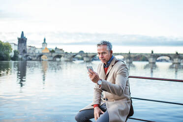 Mature handsome businessman with smartphone sitting by river Vltava in Prague city. Copy space. - HPIF23176