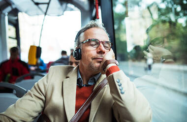 Reifer, müder Geschäftsmann mit Brille und Telefon, der mit dem Bus in der Stadt unterwegs ist und Musik hört. - HPIF23187