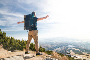 Rear view of man with backpack hiking in mountains in summer, resting with arms stretched. - HPIF23246