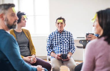 Männer und Frauen sitzen während einer Gruppentherapie im Kreis und kleben Zettel mit negativen Emotionen auf die Stirn. - HPIF23335