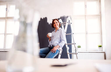Porträt einer jungen, kreativen Frau mit einer Tasse Kaffee, die die Wand schwarz anmalt, ein Startup-Unternehmen. - HPIF23375