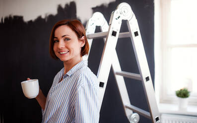 Porträt einer jungen, kreativen Frau mit einer Tasse Kaffee, die die Wand schwarz anmalt, ein Startup-Unternehmen. - HPIF23376