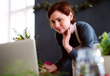 Eine junge kreative Frau arrangiert Blumen in einem Blumenladen mit einem Laptop. - HPIF23449