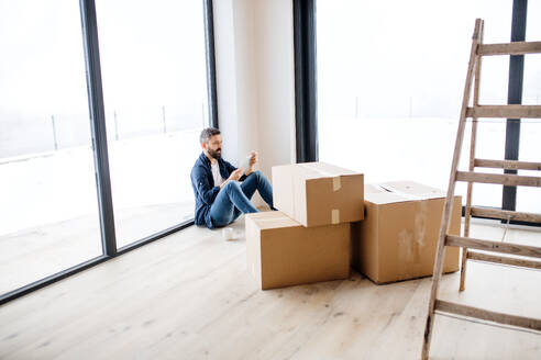 Ein reifer Mann mit Tablet und Kartons auf dem Boden sitzend, die Einrichtung eines neuen Hauses. Ein neues Haus Konzept. - HPIF23479