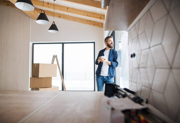 A mature man with cardboard boxes and cup of coffee standing by a window, furnishing new house. A new home concept. Copy space. - HPIF23487