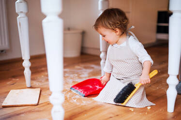 A small toddler girl with brush and dustpan sweeping messy floor in the kitchen at home. - HPIF23603
