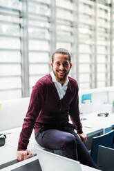 A portrait of cheerful young businessman sitting on a desk in office, looking at camera. - HPIF23653