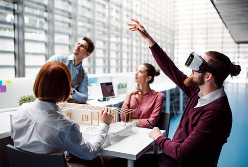 Eine Gruppe junger Architekten mit dem Modell eines Hauses und einer VR-Brille bei der Arbeit in einem Büro, im Gespräch. - HPIF23661