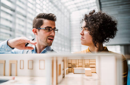 Zwei junge Architekten mit dem Modell eines Hauses stehen in einem Büro, arbeiten und unterhalten sich. - HPIF23681