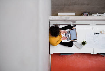 Draufsicht auf eine junge Geschäftsfrau oder Architektin, die in einem Büro auf dem Schreibtisch sitzt und einen Laptop benutzt, Kopierraum. - HPIF23683
