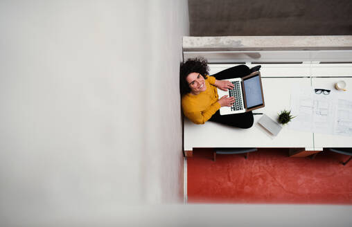 Draufsicht auf eine junge Geschäftsfrau oder Architektin, die in einem Büro auf dem Schreibtisch sitzt und einen Laptop benutzt, Kopierraum. - HPIF23684