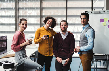 A group of cheerful young businesspeople with cup of coffee standing in office, looking at camera. - HPIF23687