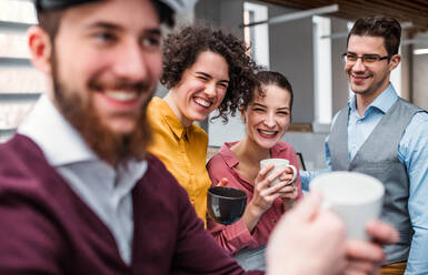 A group of cheerful young businesspeople with cup of coffee standing in office, laughing. - HPIF23689