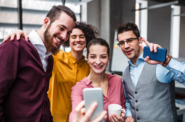 Eine Gruppe junger Berufstätiger im Büro, die einen Moment der Freude mit einem Selfie mit ihren Smartphones festhalten - HPIF23692