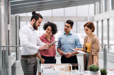 Eine Gruppe junger Architekten mit einem Tablet und dem Modell eines Hauses steht in einem Büro und arbeitet und spricht. - HPIF23713