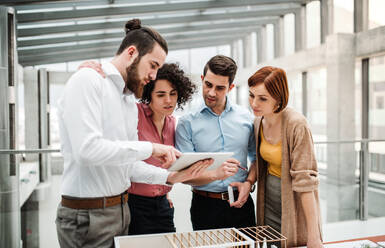 Eine Gruppe junger Architekten mit einem Tablet und dem Modell eines Hauses steht in einem Büro und arbeitet und spricht. - HPIF23714