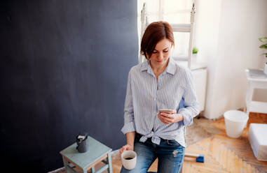 A portrait of young creative woman with a cup of coffee and smartphone painting wall black. A startup of small business. - HPIF23730
