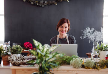 Eine junge kreative Frau arrangiert Blumen in einem Blumenladen mit einem Laptop. - HPIF23750