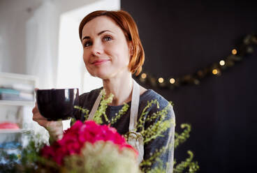 Eine junge, kreative Frau mit einem Kaffee, die in einem Blumenladen Blumen arrangiert, eine Neugründung eines Blumenladens. - HPIF23758