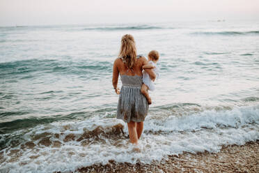 Rückansicht einer jungen Mutter mit einem Kleinkind, das im Sommerurlaub am Meer spazieren geht. - HPIF23778