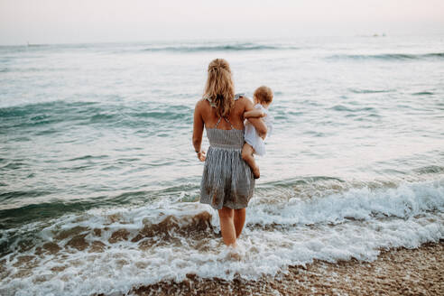 Rückansicht einer jungen Mutter mit einem Kleinkind, das im Sommerurlaub am Meer spazieren geht. - HPIF23778