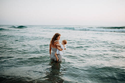 Rückansicht einer jungen Mutter mit einem Kleinkind, das im Sommerurlaub am Meer spazieren geht. - HPIF23779