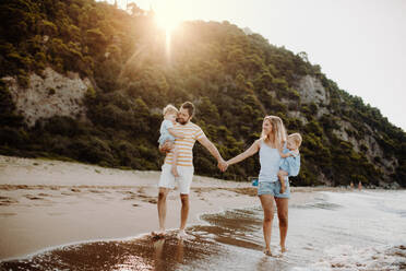 Eine Familie mit zwei Kleinkindern, die im Sommerurlaub bei Sonnenuntergang am Strand spazieren gehen. Ein Vater und eine Mutter tragen Sohn und Tochter in den Armen und halten sich an den Händen. - HPIF23795